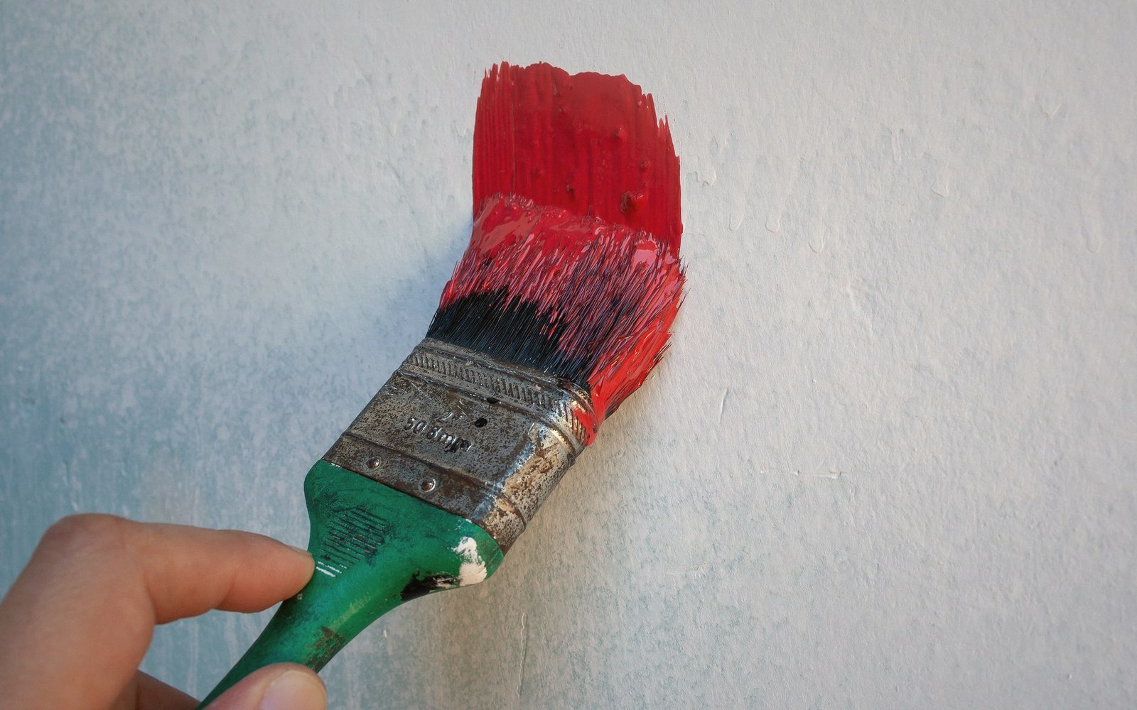 A hand uses a paintbrush to apply red paint onto a textured wall