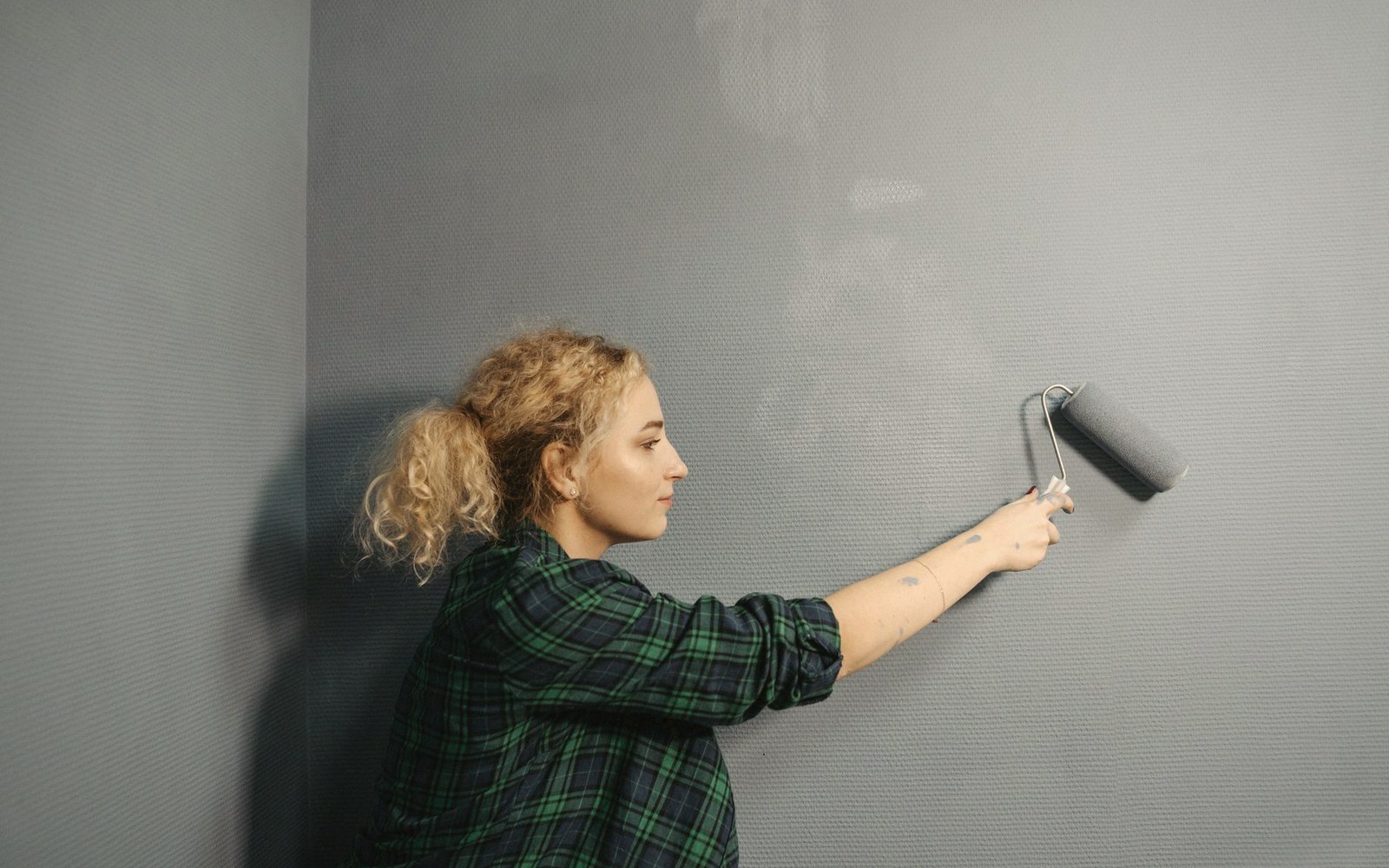 Woman in a plaid shirt uses a roller to paint a wall grey