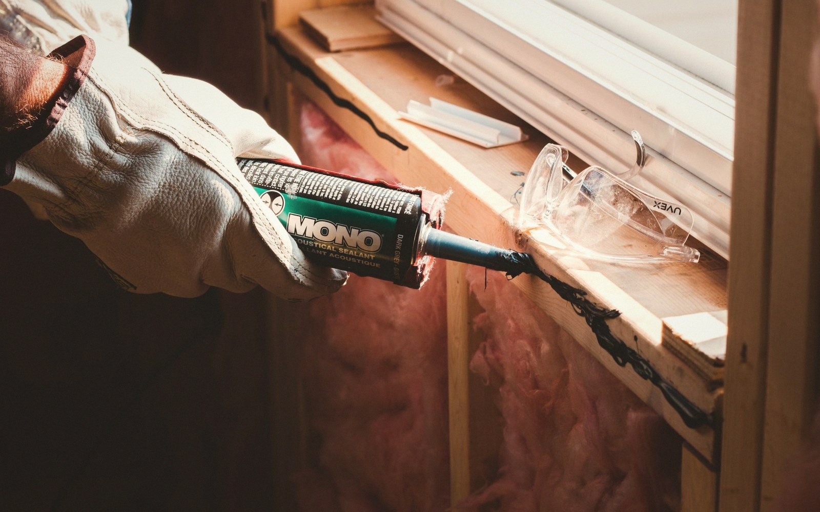 Gloved hand applying acoustical sealant along a window frame near pink insulation