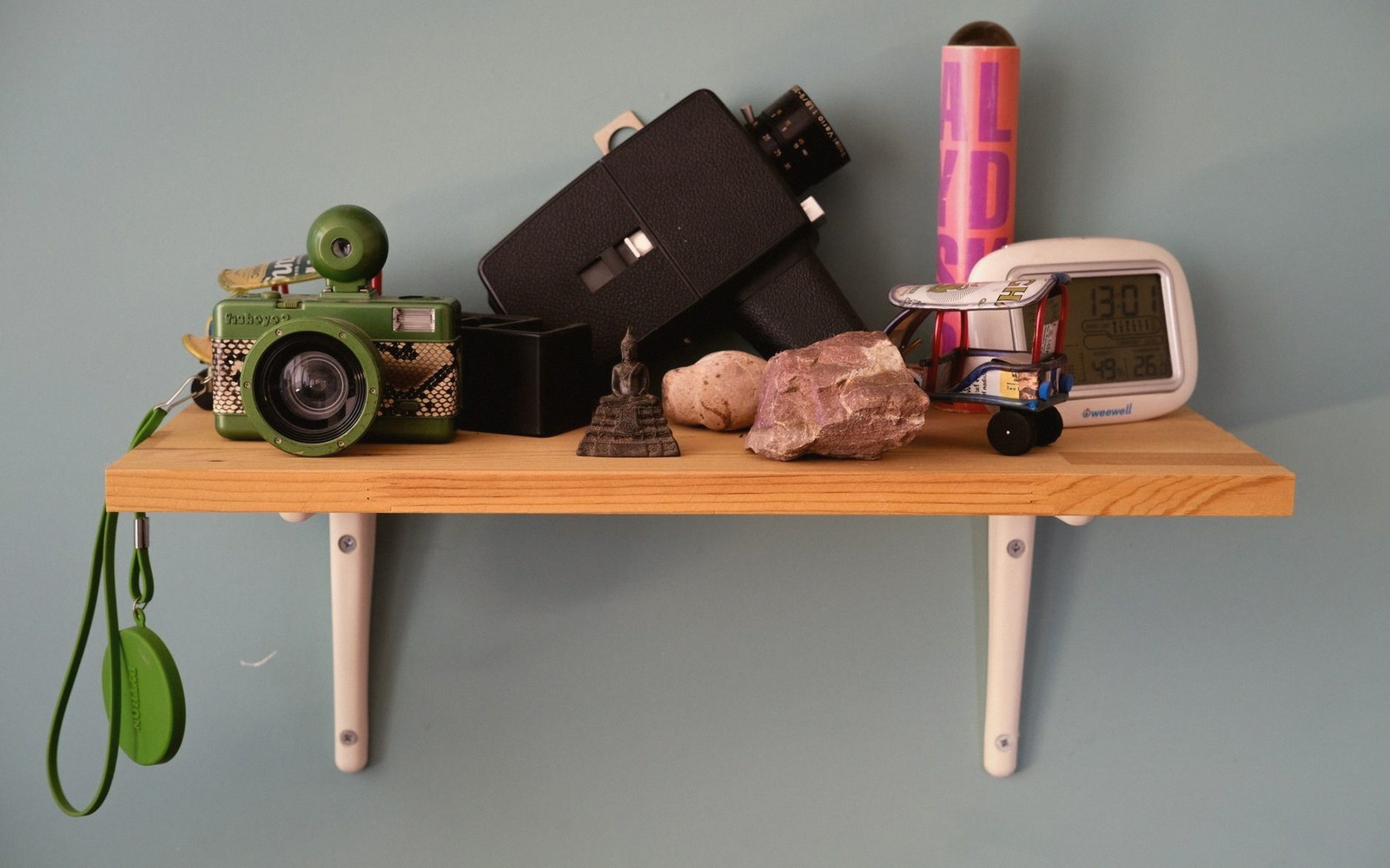 A wooden shelf holds a toy camera, vintage movie camera, small statue, rocks, and a digital clock