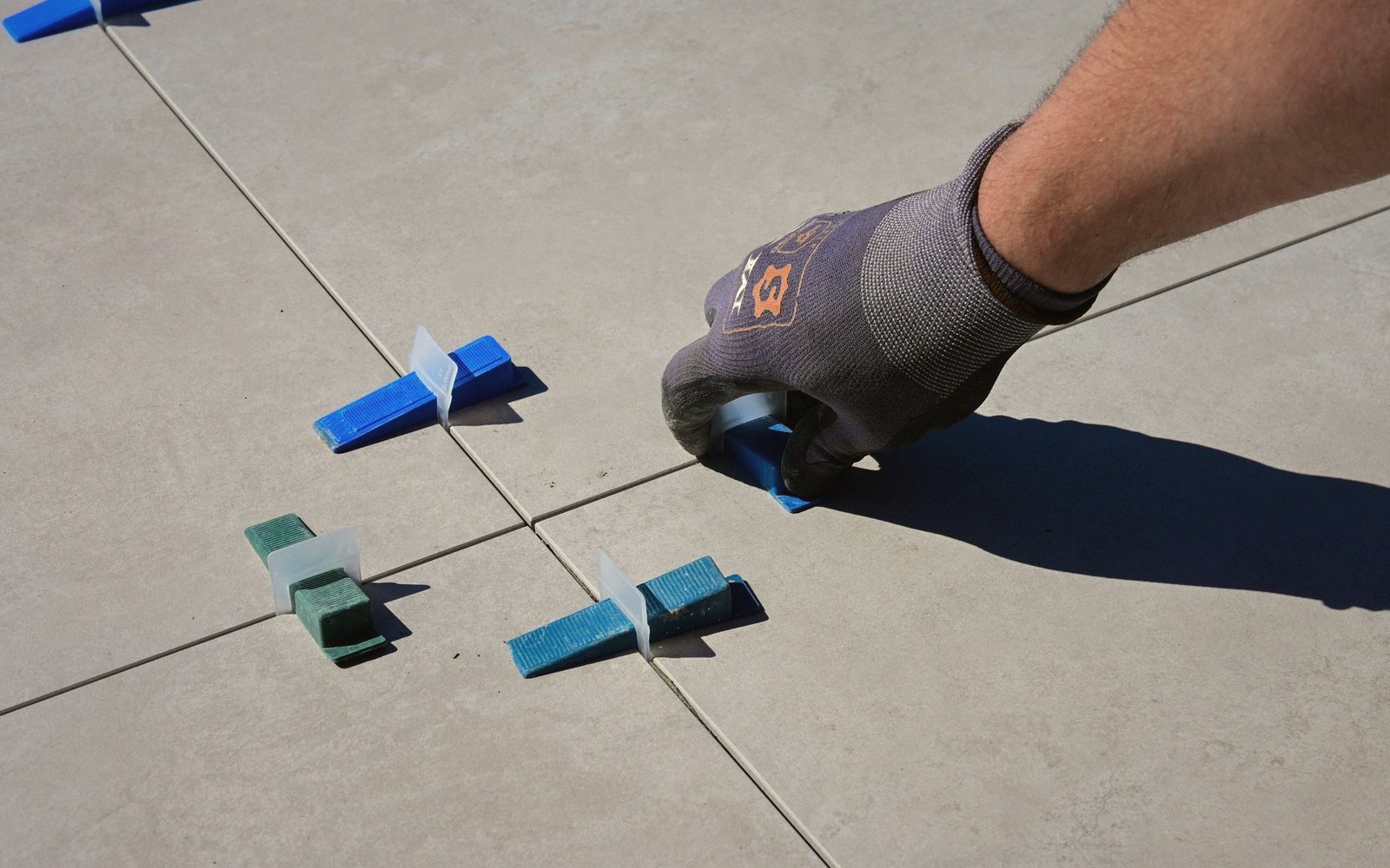 A gloved hand tightens a plastic tile leveling wedge between floor tiles