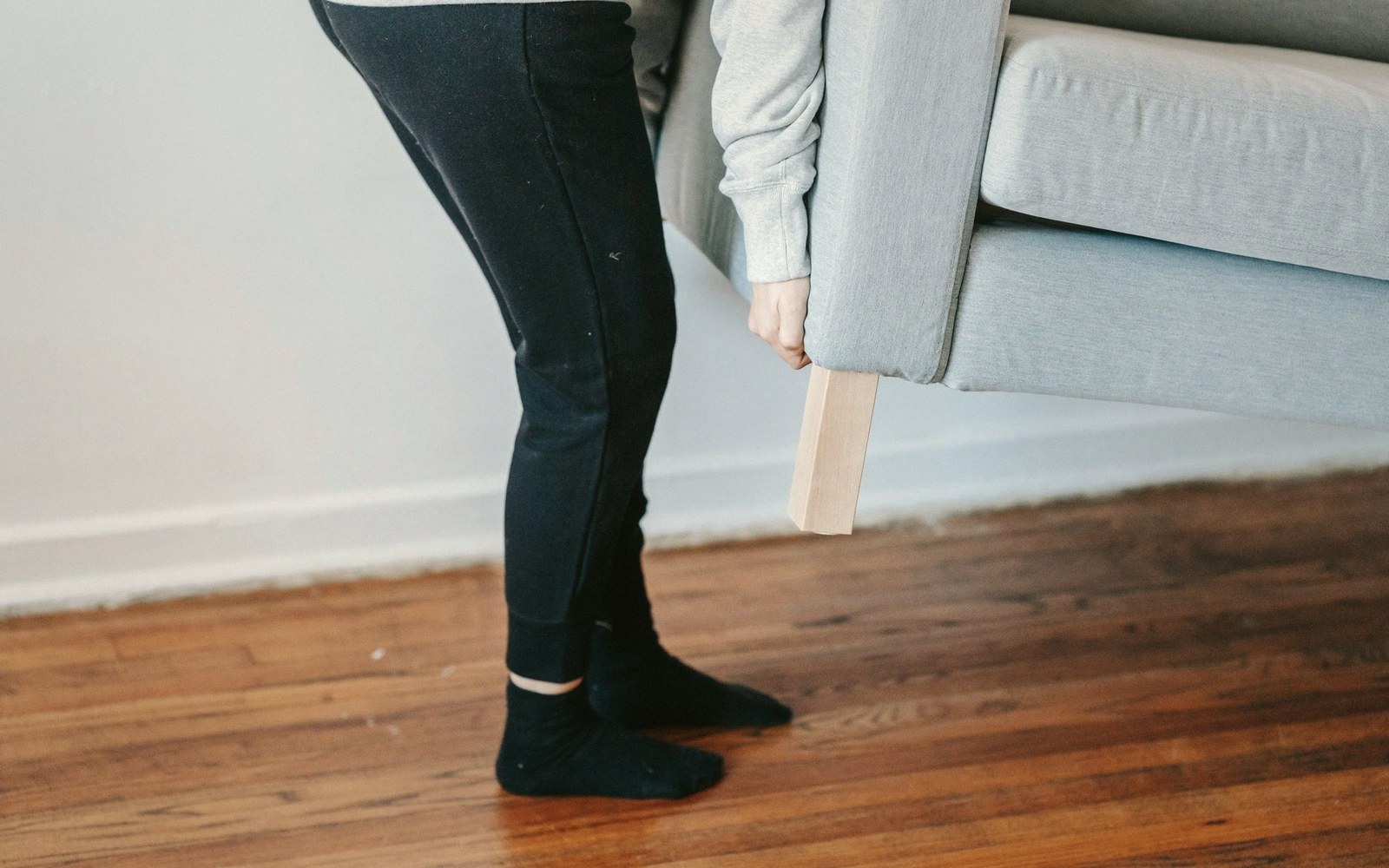 A person tilts a gray sofa on a wooden floor to check its wooden leg
